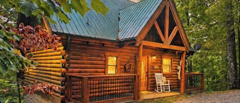 A log cabin with a metal roof, a porch with a chair, and a gravel driveway.