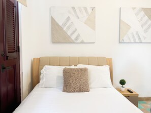 Ein ordentlich bezogenes Bett mit weißen Leinen, einem beigen Kopfteil und einem strukturierten, beigen Kissen. Zwei abstrakte Kunstwerke hängen an der Wand darüber.