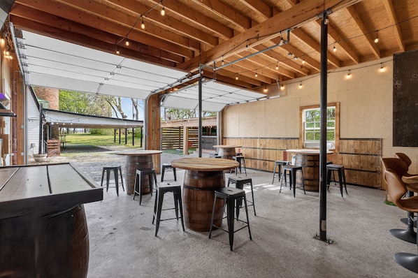 An outdoor seating area with wooden tables and stools, a covered section with a view of the yard, and a rustic interior with exposed beams and a concrete floor.