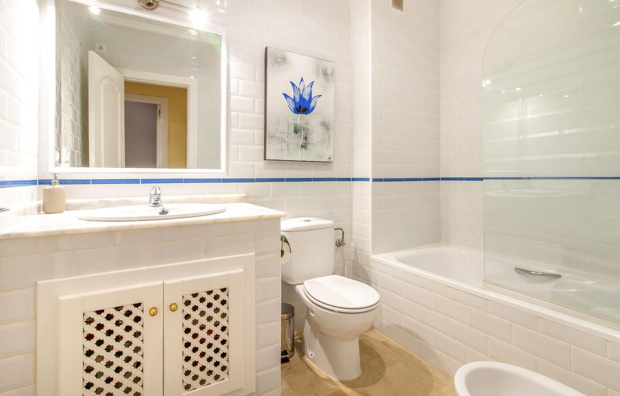 Ein Badezimmer mit weißem Waschbecken, WC und Badewanne. Über dem Waschbecken befindet sich ein Spiegel und an der Wand ein blaues Blumenbild.