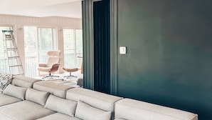 Ein modernes Wohnzimmer mit einer Eckcouch, einer dunkel abgesetzten Wand und Blick ins Freie durch große Fenster.