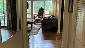 Un pasillo que da a una sala de estar con suelo de madera, ventanas amplias, una silla, un sofá y una lámpara.