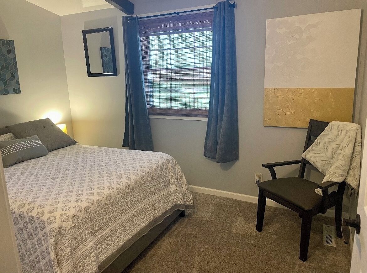 A bedroom with a bed, a chair, a window with curtains, a wall-mounted mirror, and a painting.