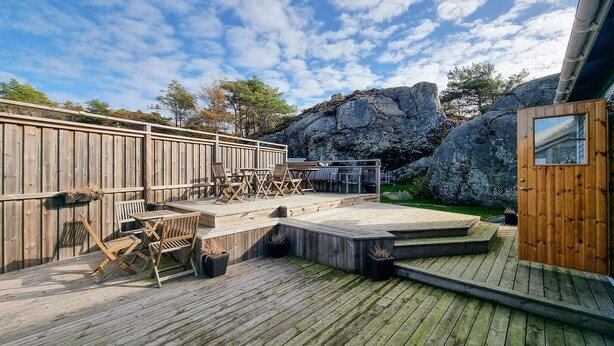 En træterrasse med udendørs møbler, en klippeformation og en træbygning med et vindue.