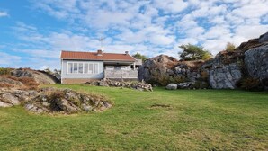 Et lille hus med røde tegltag, hvide vægge og en træterrasse. Det er omgivet af en græsplæne og store sten.