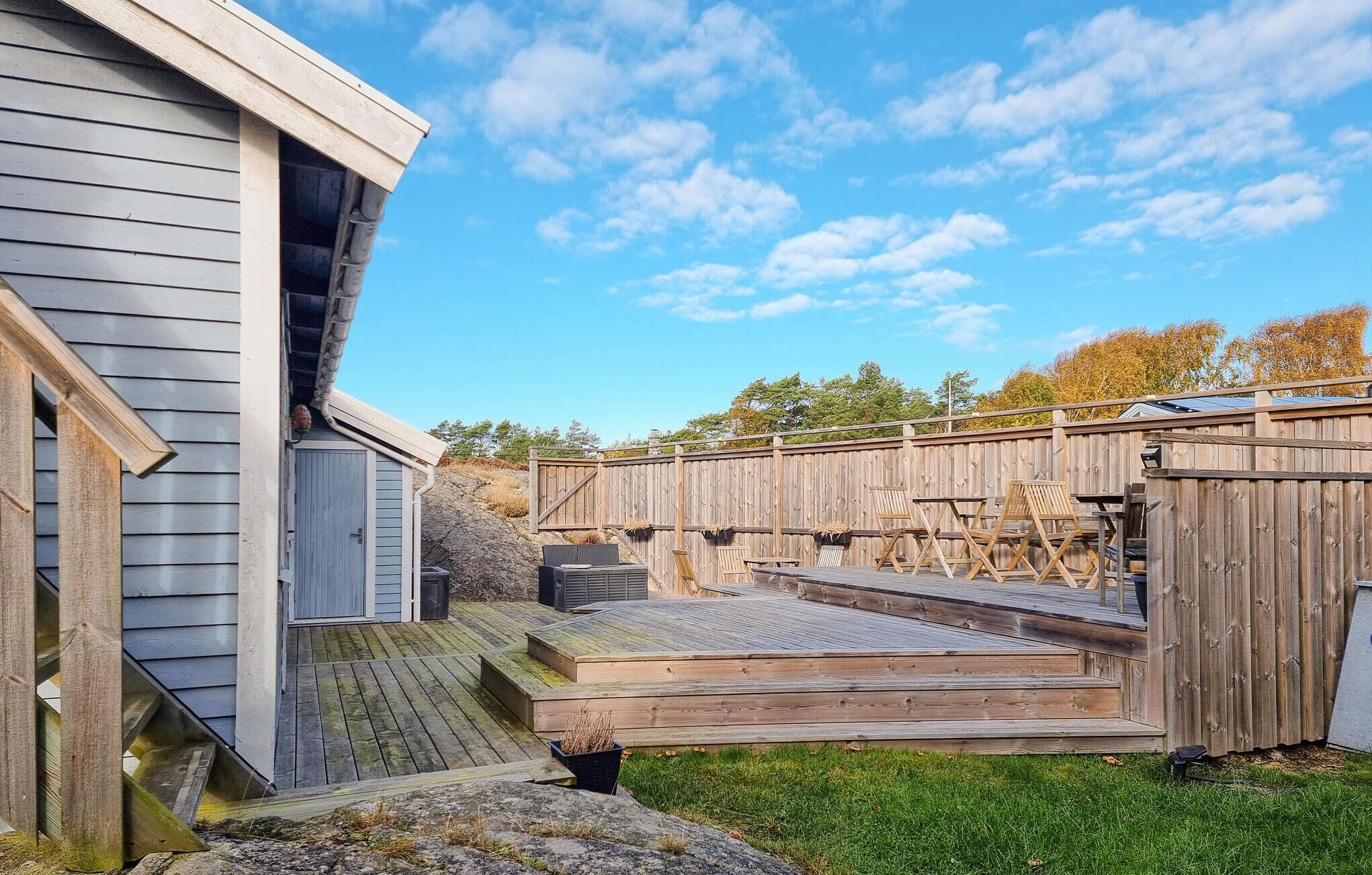 Un patio in legno con posti a sedere all'aperto e una zona riparata, circondato da una recinzione in legno e una casa con tetto spiovente.