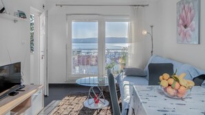 Ein modernes Wohnzimmer mit Balkon, einem Fernseher, einem Esstisch mit Obstschale und einem Bild an der Wand.