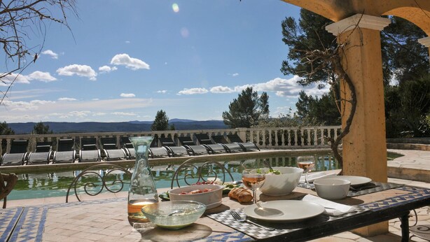 A dining table set with plates, bowls, and a bottle, overlooking a pool with lounge chairs.