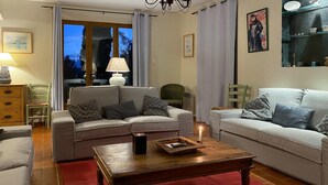 A living room with two sofas, a wooden coffee table, and a red rug. There is a window with curtains, a lamp, and a painting on the wall.