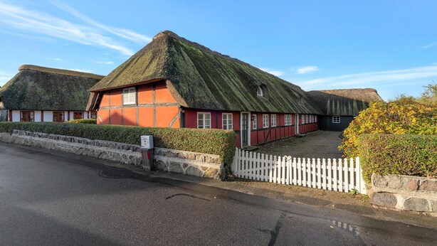 A traditional thatched-roof house with a red exterior, a white picket fence, and a stone wall.