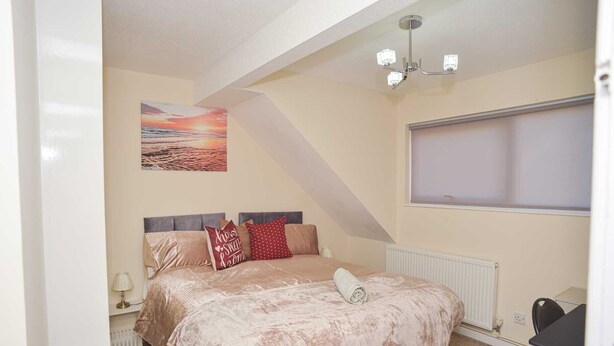 Een slaapkamer met een bed, nachtkastje, lamp, radiator, raam en een plafondlamp.
