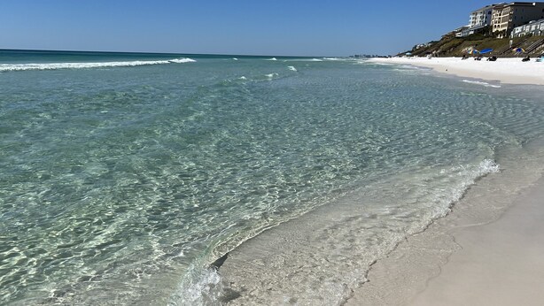 Ein Strand mit klarem, flachen Wasser, einem Sandufer und einem Gebäude mit mehreren Balkonen im Hintergrund.