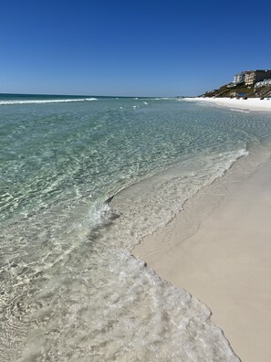 A beach with clear shallow waters, a sandy shore, and a building with multiple balconies in the background.