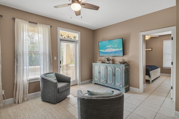 A living room with a ceiling fan, a television mounted on the wall, a cabinet, and a bed in the adjacent room.