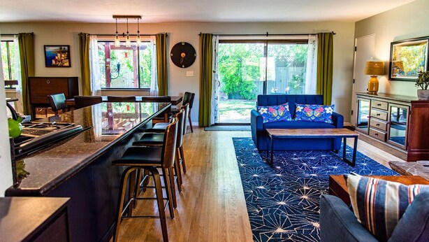 A modern kitchen with a central island, bar stools, and a dining table. There is a blue sofa with a colorful cushion, a patterned rug, and a view of greenery through large windows. - Family Home Near Bidwell Park (Chico)