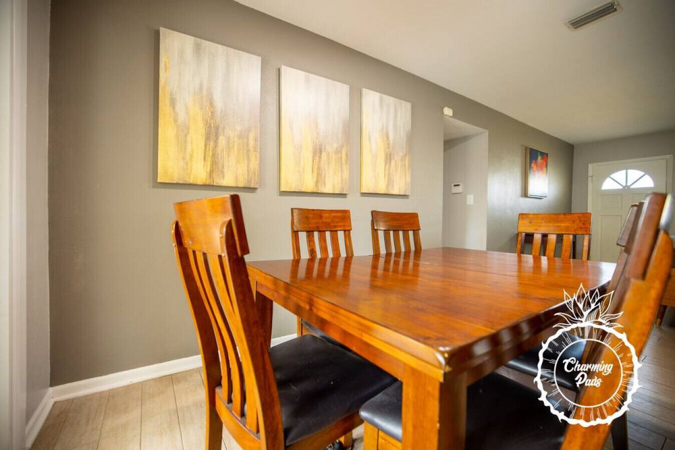 Comedor con mesa de madera y seis sillas, tres cuadros en la pared y una puerta al fondo.