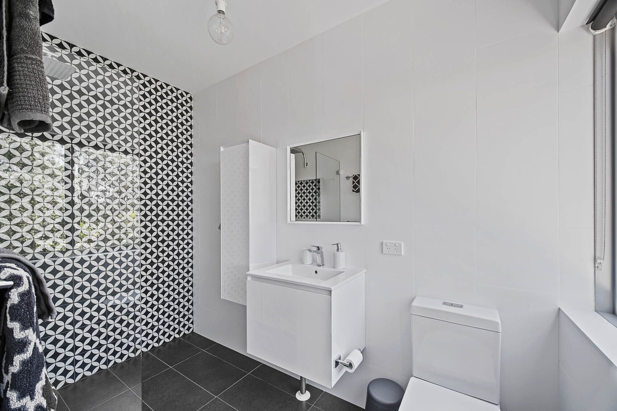 Un baño moderno con lavamanos, espejo y retrete blancos. Las paredes presentan un diseño de baldosas con motivos geométricos.