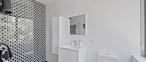 Un baño moderno con lavamanos, espejo y retrete blancos. Las paredes presentan un diseño de baldosas con motivos geométricos.
