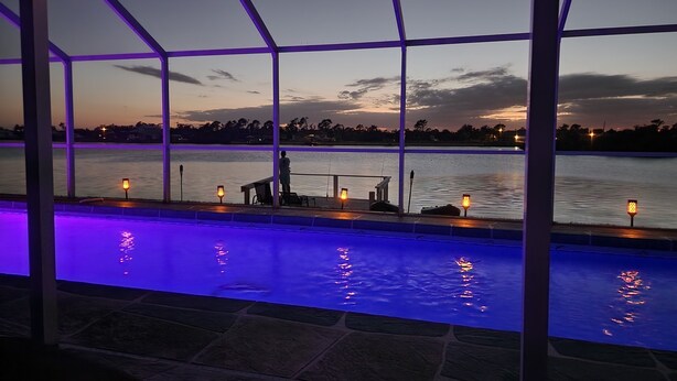 An indoor pool with a view of a lake at dusk, surrounded by a glass enclosure with purple lighting. - Tranquil Waterfront Pool Home with beautiful views and sunsets and great fishing (Port Charlotte)