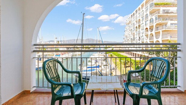 A balcony with two green chairs and a small table, overlooking a marina with boats and a building with balconies. - WELCS DUPLEX 025 EMP frente al Mar (Empuriabrava)