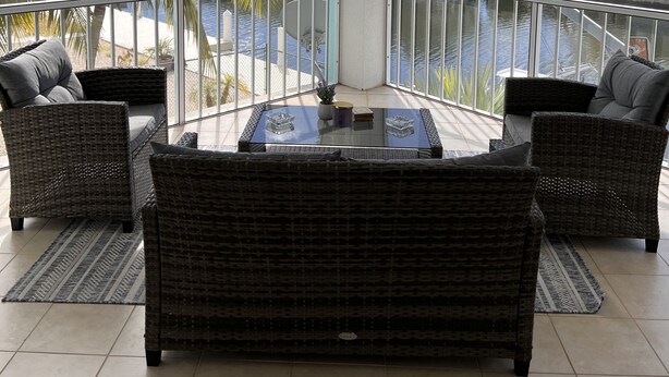 A balcony with wicker furniture, a glass table, and a view of a pool and palm trees.