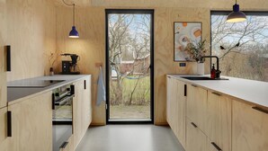Et moderne køkken med træskabe, en kaffemaskine og et stort vindue med udsigt til træer og et hus.