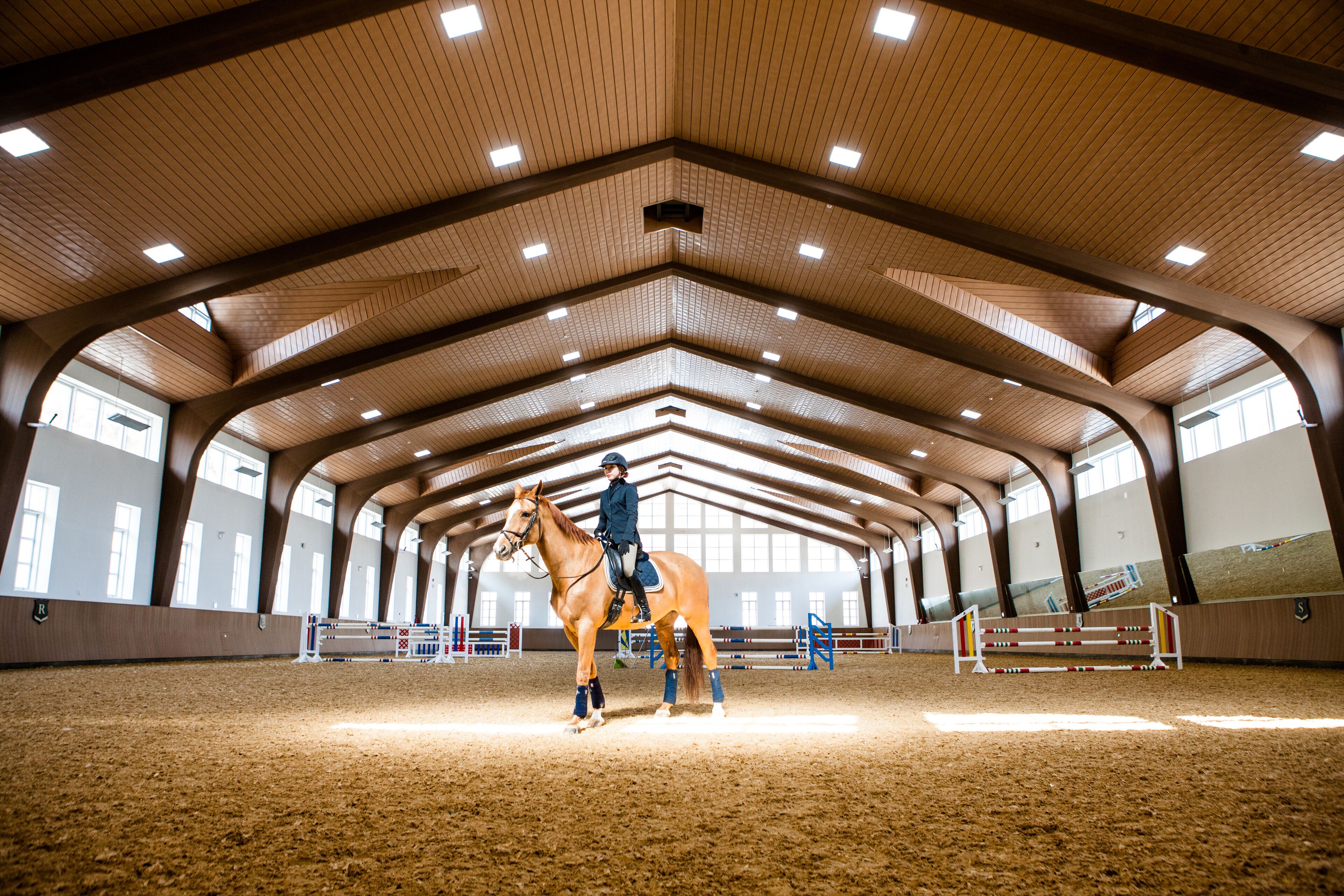 An indoor equestrian arena with a high ceiling, wooden beams, and a sandy floor.