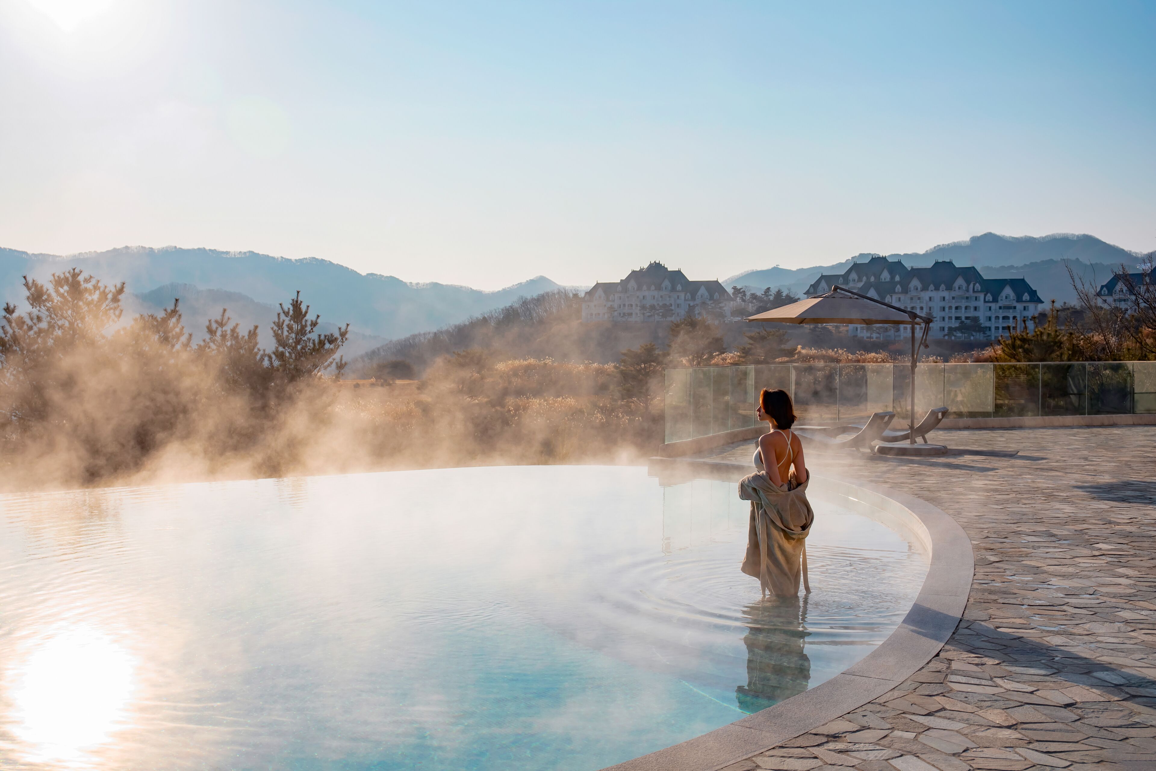 A spa pool with mist, a person in a robe, and a mountainous backdrop.