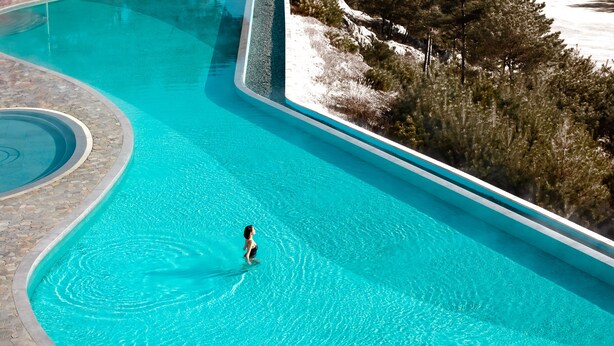 A large, clear swimming pool with a person swimming, surrounded by a stone deck and trees. - Sono Felice Village Vivaldi Park (Hongcheon)