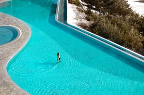 A large, clear swimming pool with a person swimming, surrounded by a stone deck and trees.