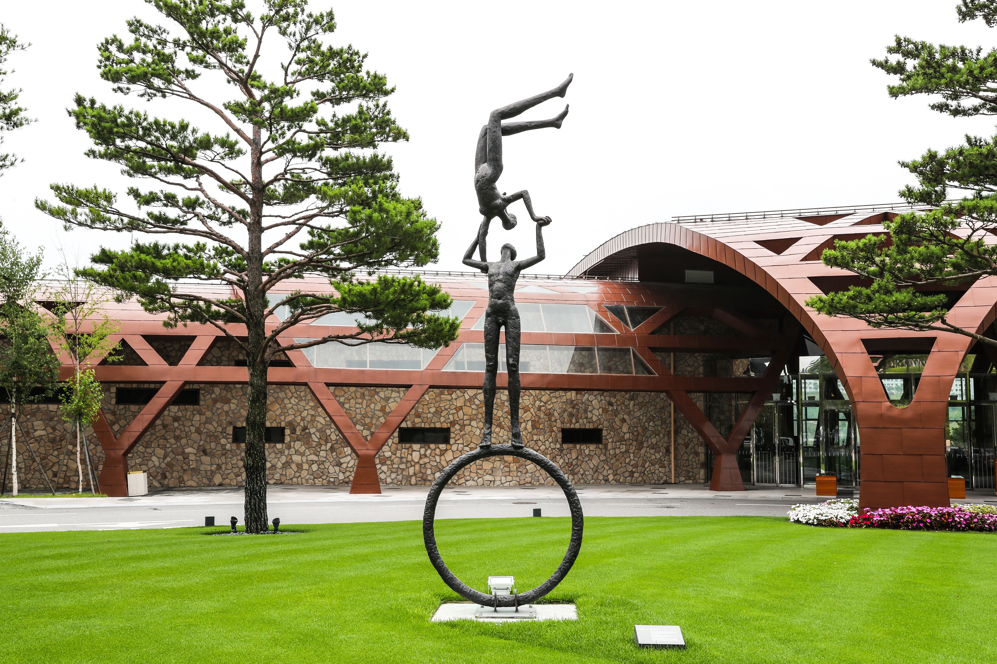 Una escultura de un hombre equilibrándose sobre un aro, ubicada en un área cubierta de césped con un edificio moderno al fondo.