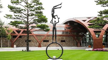 Una escultura de un hombre equilibrándose sobre un aro, ubicada en un área cubierta de césped con un edificio moderno al fondo.