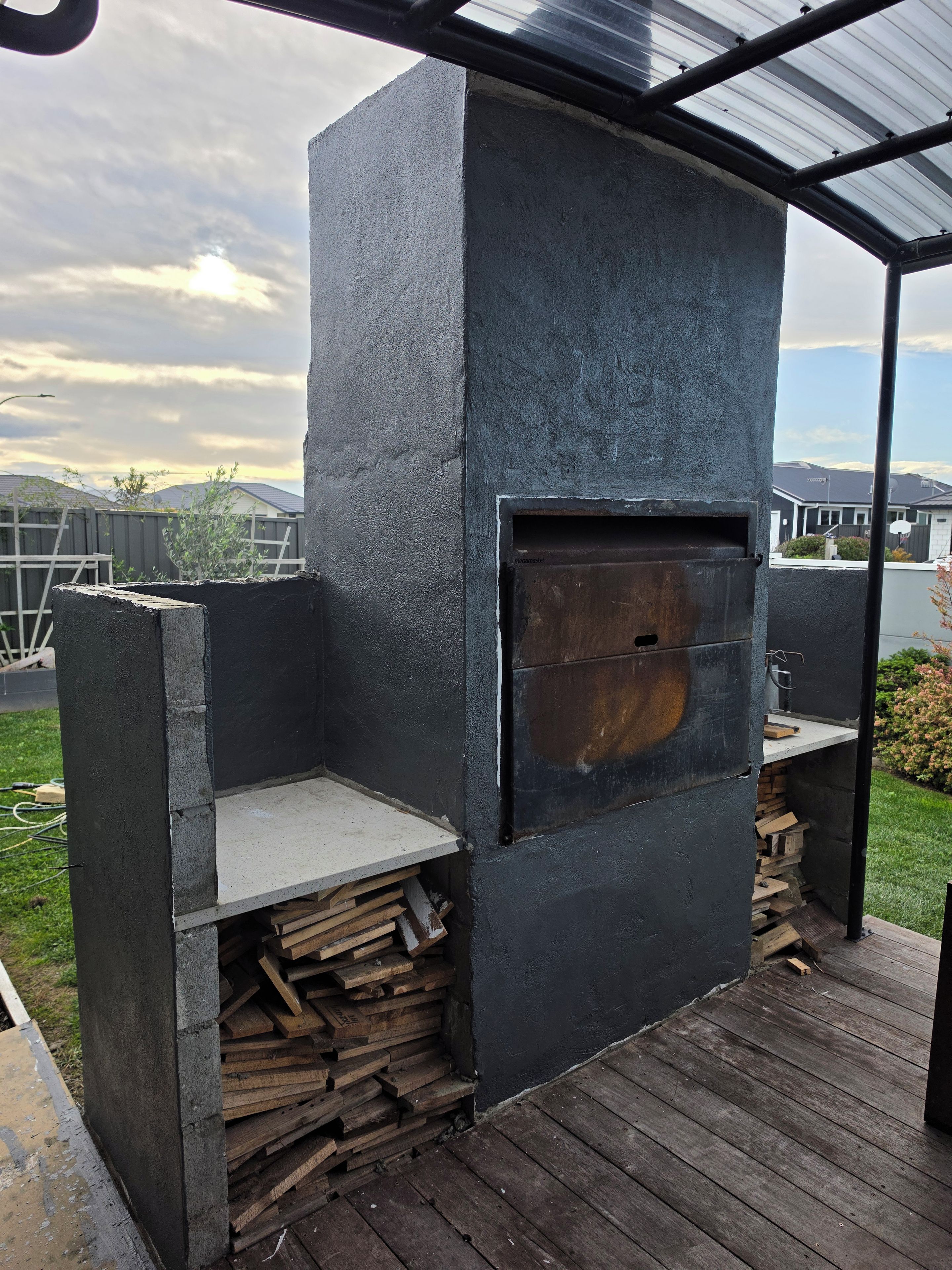 A metal outdoor grill with a built-in oven, a shelf for wood, and a stack of firewood.