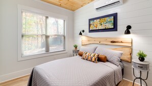 Un dormitorio con techo de madera, paredes blancas, una cama grande con colcha acolchada, cabecera de madera, una ventana con persianas, un equipo de aire acondicionado, una lámpara de pared y una mesita con una planta.
