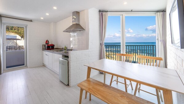 A modern kitchen with a view of the sea, a dining area with a wooden table and bench, and a sliding glass door leading to an outdoor balcony. - Gosung Stager (Goseong)