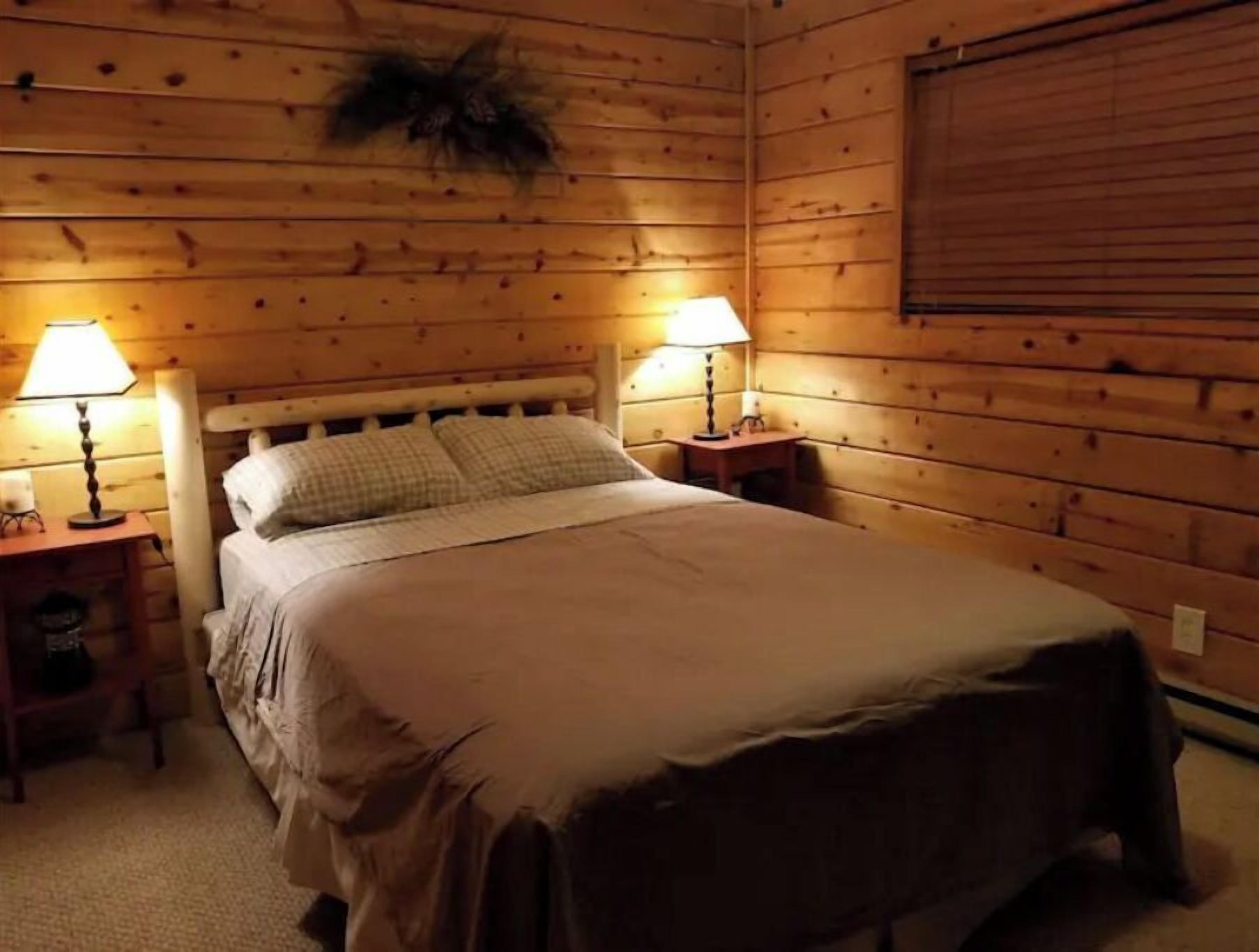 A wooden cabin room with a bed, two bedside lamps, and a window with blinds.