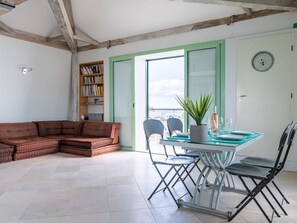 Uma sala de estar com sofá seccional, uma mesa de jantar com cadeiras, uma estante e vista através de uma porta de vidro.