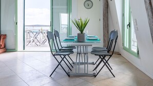 Comedor con una mesa preparada para cuatro, rodeado de sillas, una planta en maceta y vistas al mar a través de puertas abiertas.