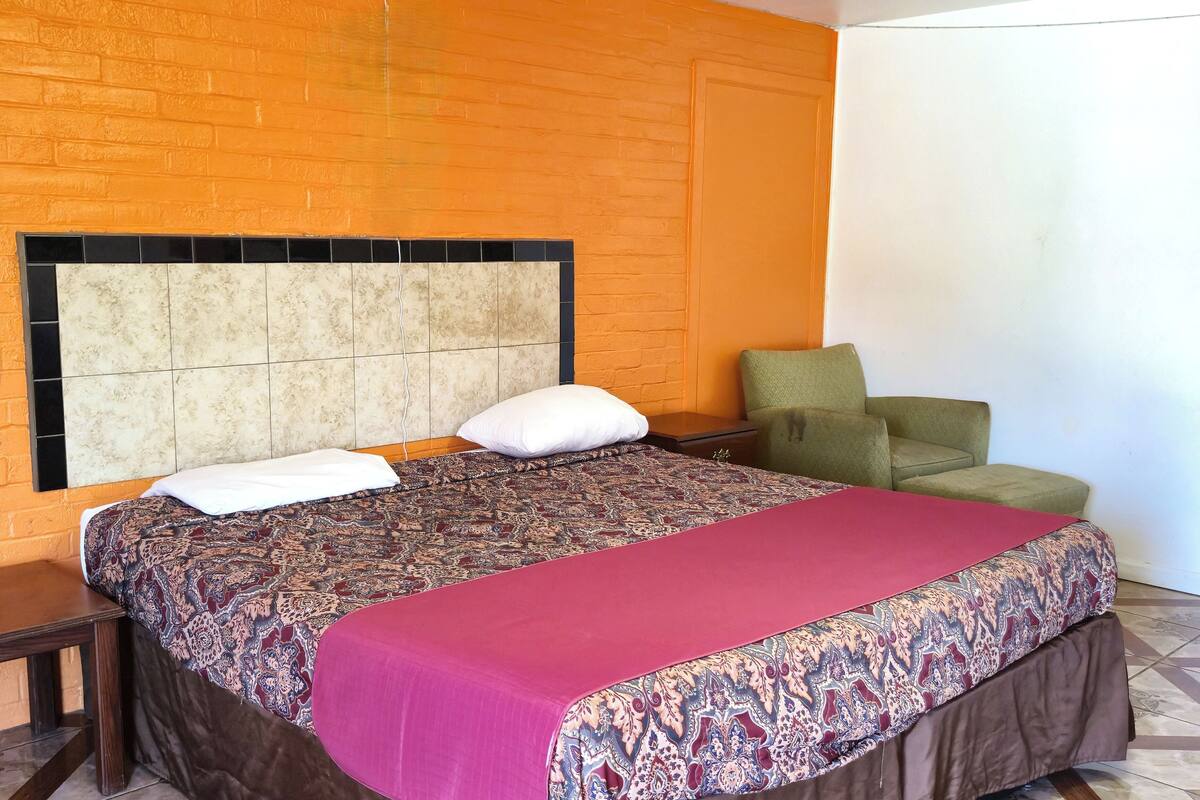 A hotel room with a patterned bedspread, a red blanket, a headboard with a tiled design, a green armchair, and a white door.