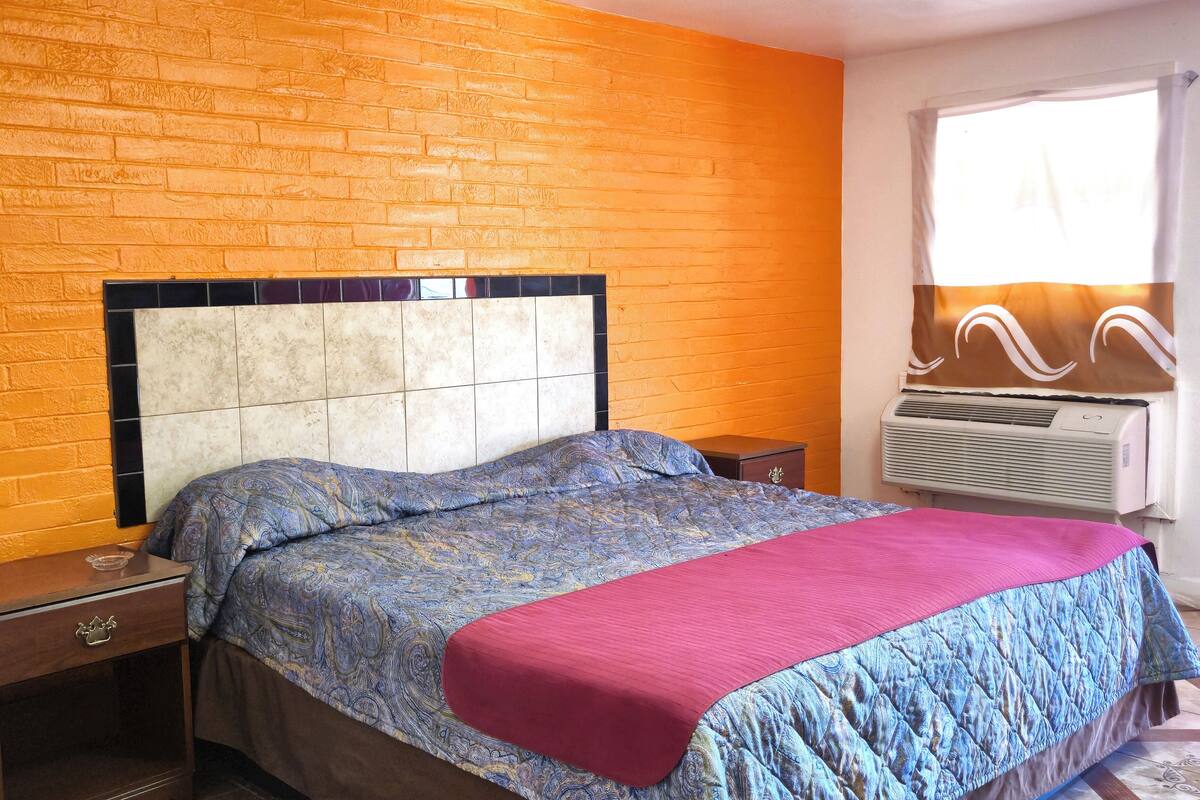 A bedroom with a brick wall, a bed with patterned bedding, a bedside table, and an air conditioning unit.