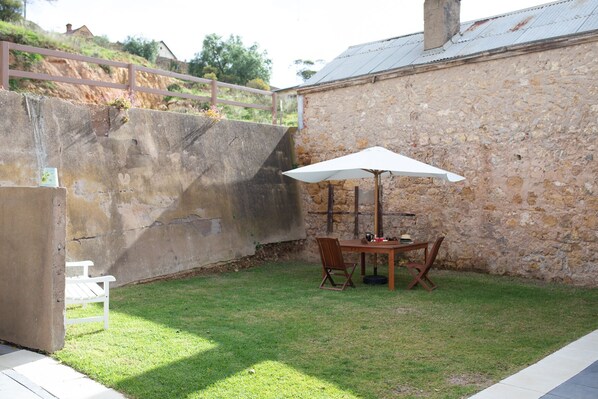 Un muro de piedra, una silla blanca, una mesa con sillas y una sombrilla en un espacio al aire libre cubierto de césped.