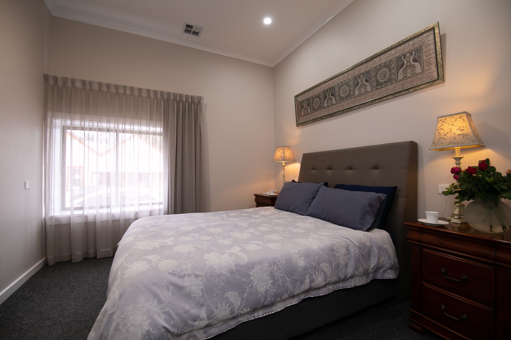 A bedroom with a large bed, two bedside tables with lamps, a decorative wall piece, and a window with curtains.