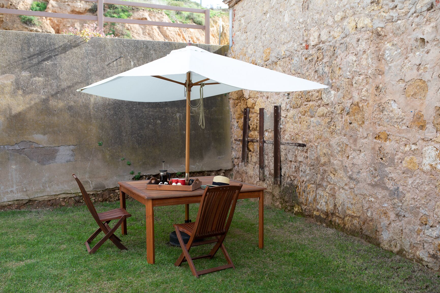 A wooden table with a white umbrella, two wooden chairs, and a tray with a coffee pot and cups.