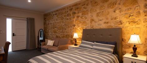 A bedroom with a stone wall, a bed with striped bedding, two bedside tables with lamps, a sofa, and a door.