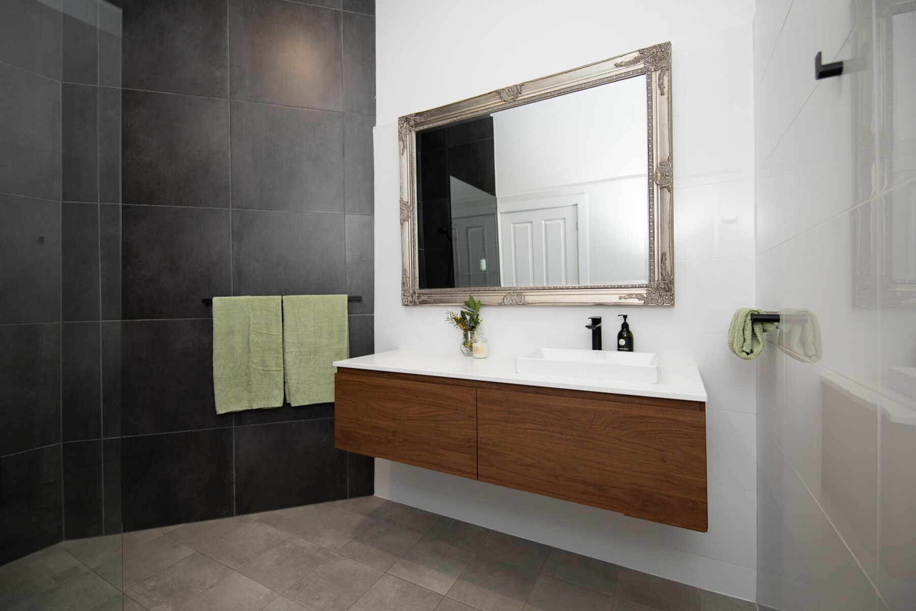 A bathroom with a white vanity, dark tiled walls, and a large mirror with an ornate frame.