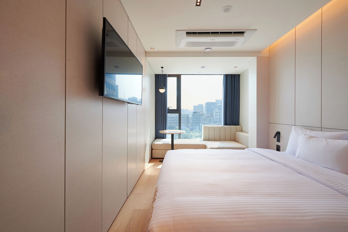 A modern hotel room with a large bed, a flat-screen TV mounted on the wall, a small round table, and a view of the city through large windows.