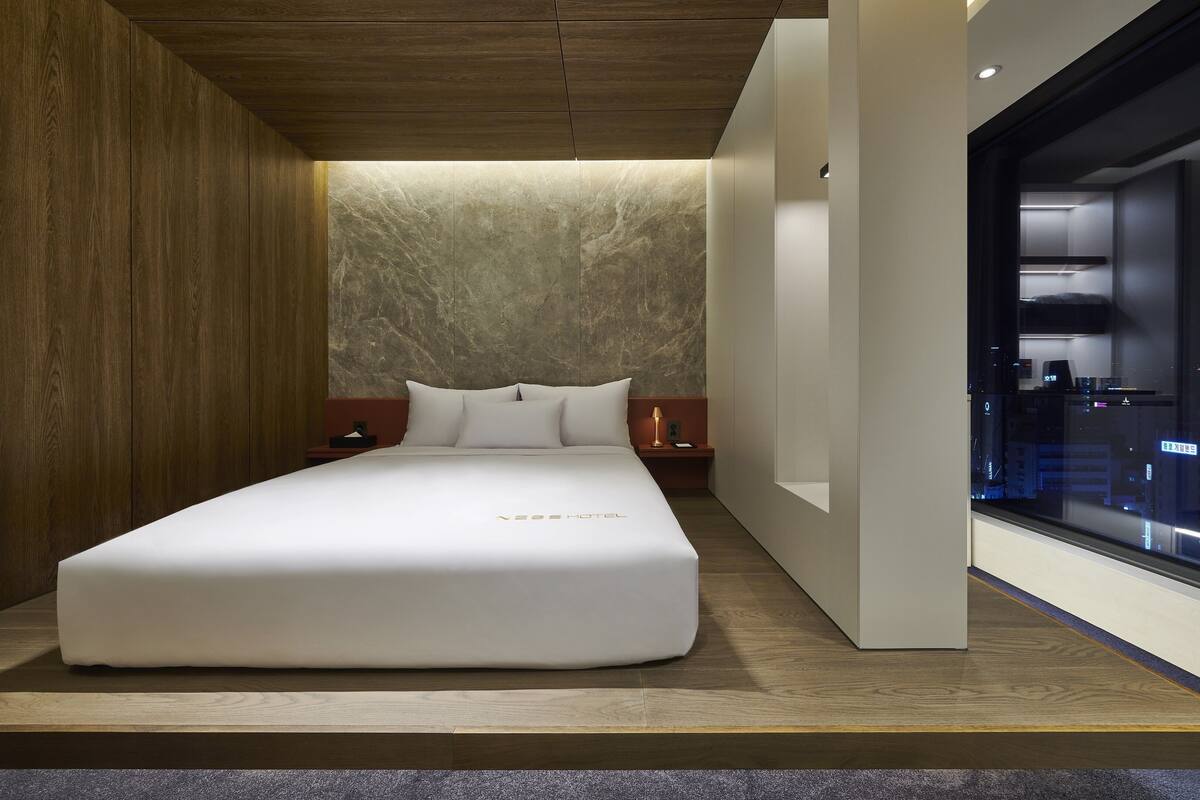 A modern hotel room with a large bed, a marble wall, and a city view through a window.