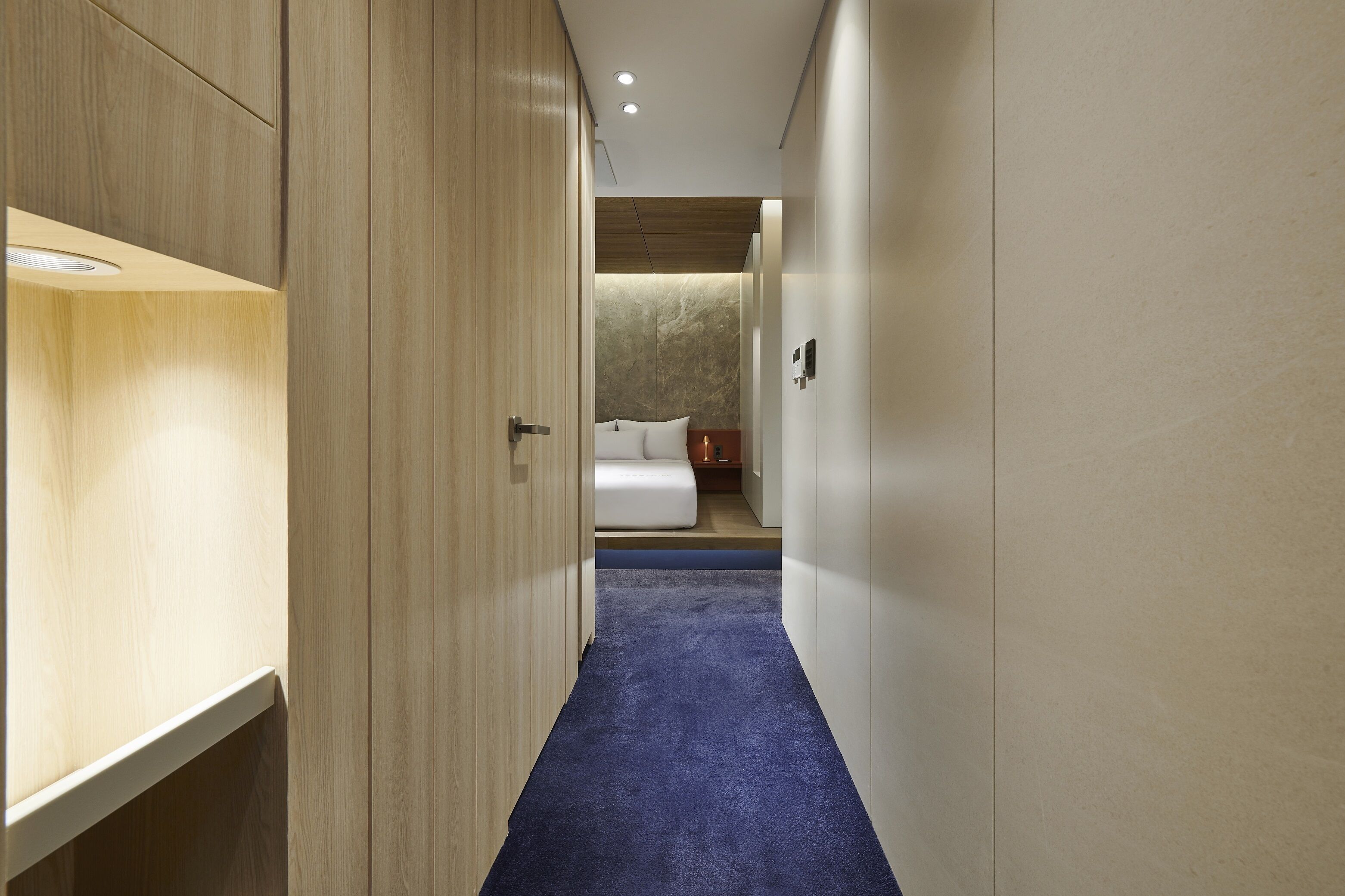 A hallway leading to a bedroom with a bed, wooden wardrobe, and a blue carpet.