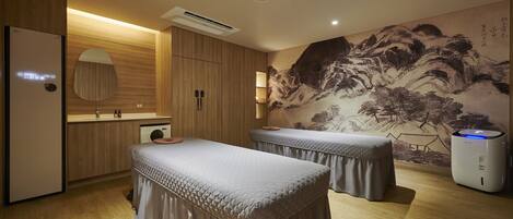 A room with two massage tables, wooden flooring, a wall-mounted air conditioner, and a traditional painting.