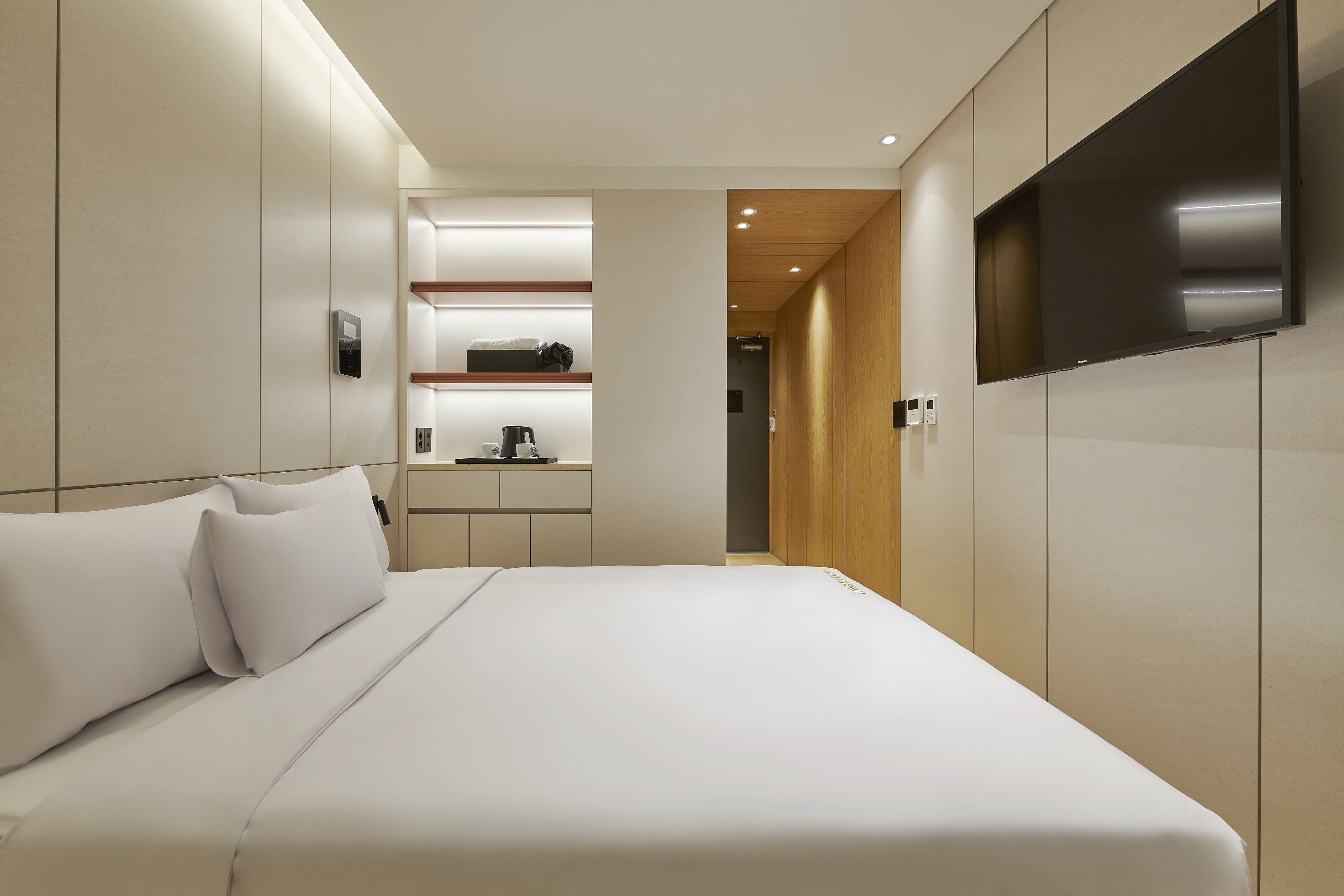 A modern hotel room with a large bed, a flat-screen TV mounted on the wall, a built-in shelf with a coffee machine, and a wooden door leading to another room.